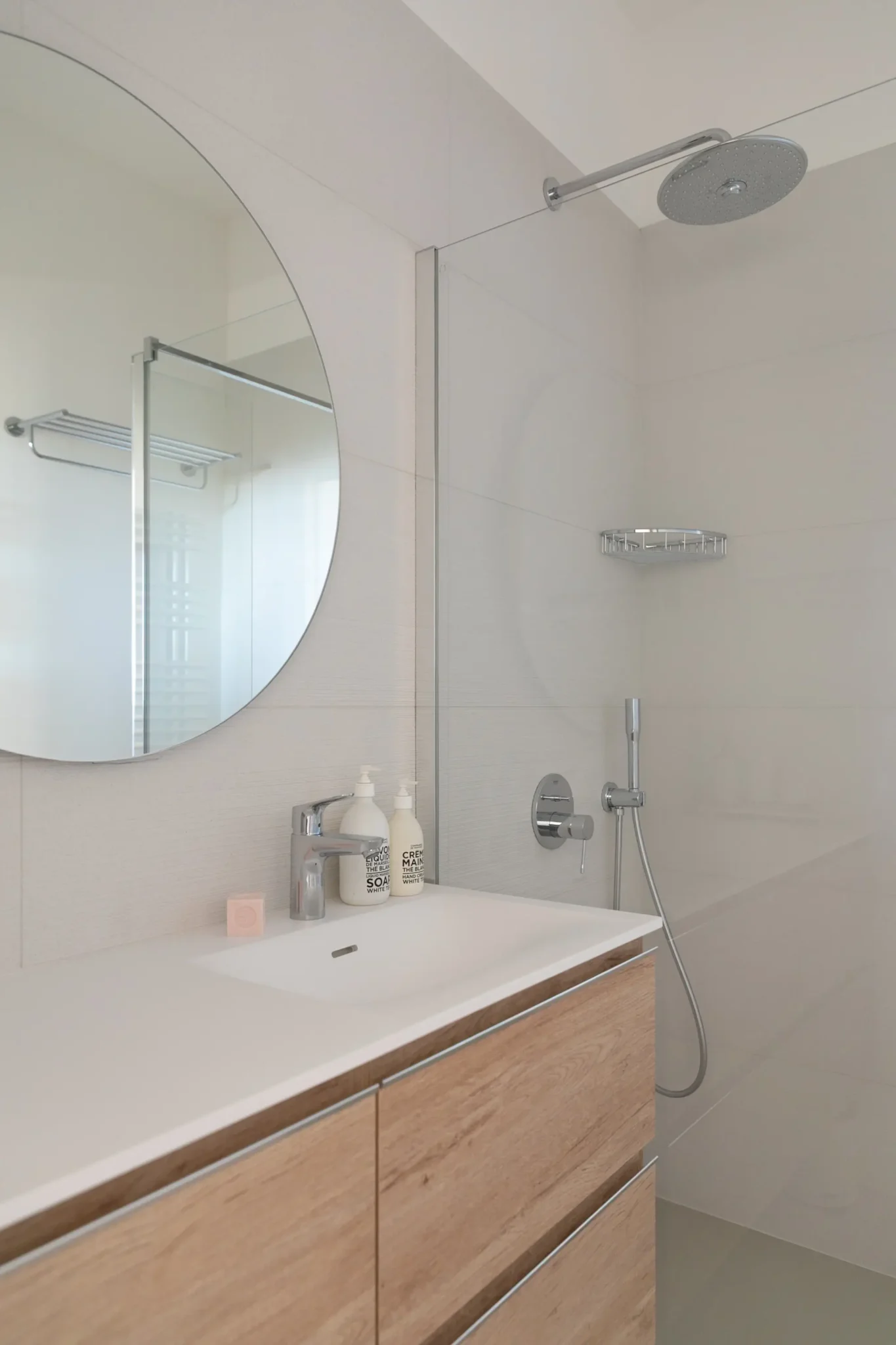 salle de bains avec douche à l'italienne paroi vitrée et ciel de pluie miroir rond meuble vasque avec tiroirs façade bois robinetterie inox